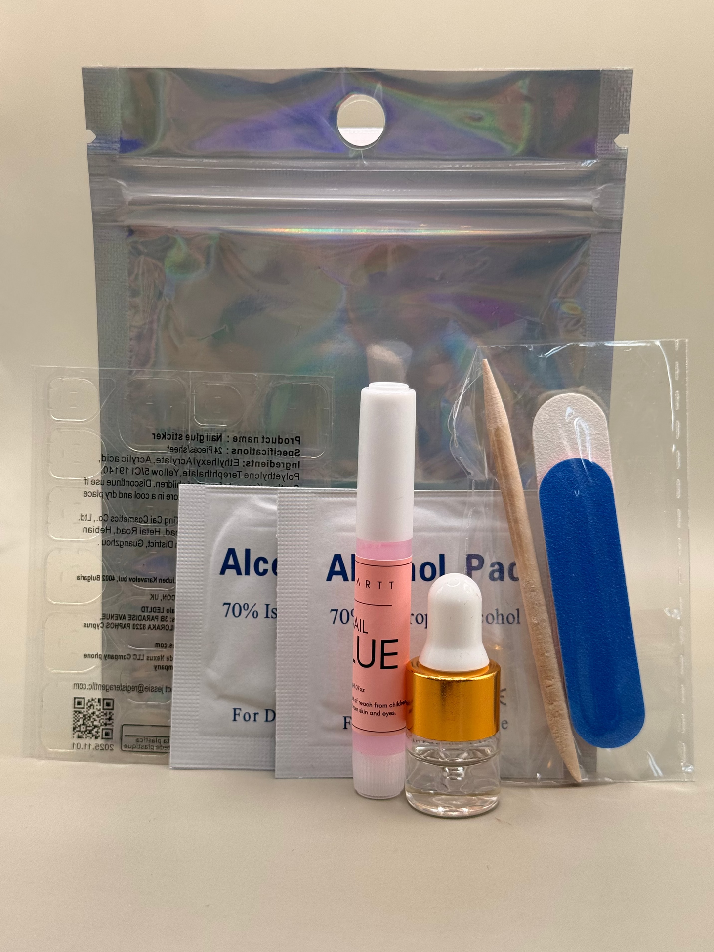 A beauty kit containing a small glass dropper bottle with a gold collar, a pink lip gloss tube, a clear plastic packet with blue text, a wooden spatula, a blue nail file, and an iridescent zip pouch on a light beige background.