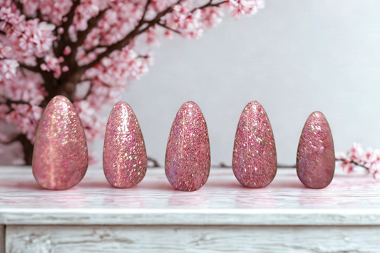 Five pink glittery almond shaped nails on a white surface with cherry blossom branches in the background.