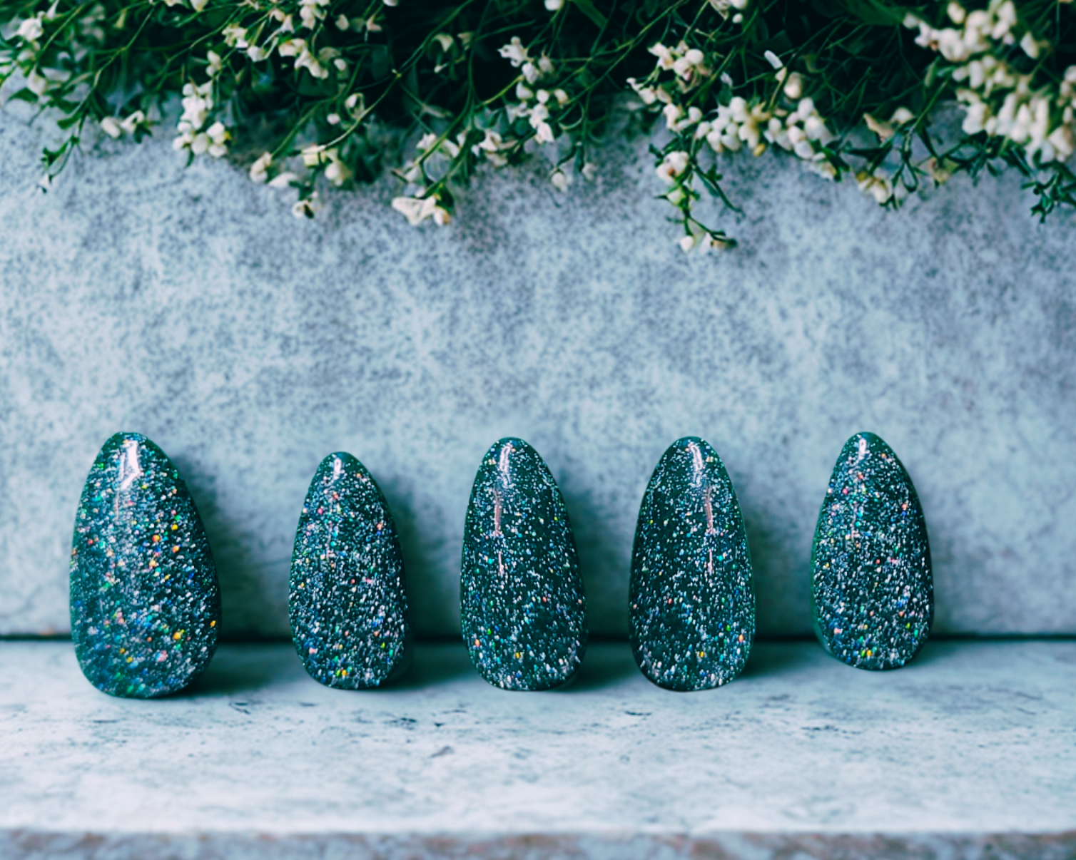 Five green glittery almond nails on a stone surface with a floral background